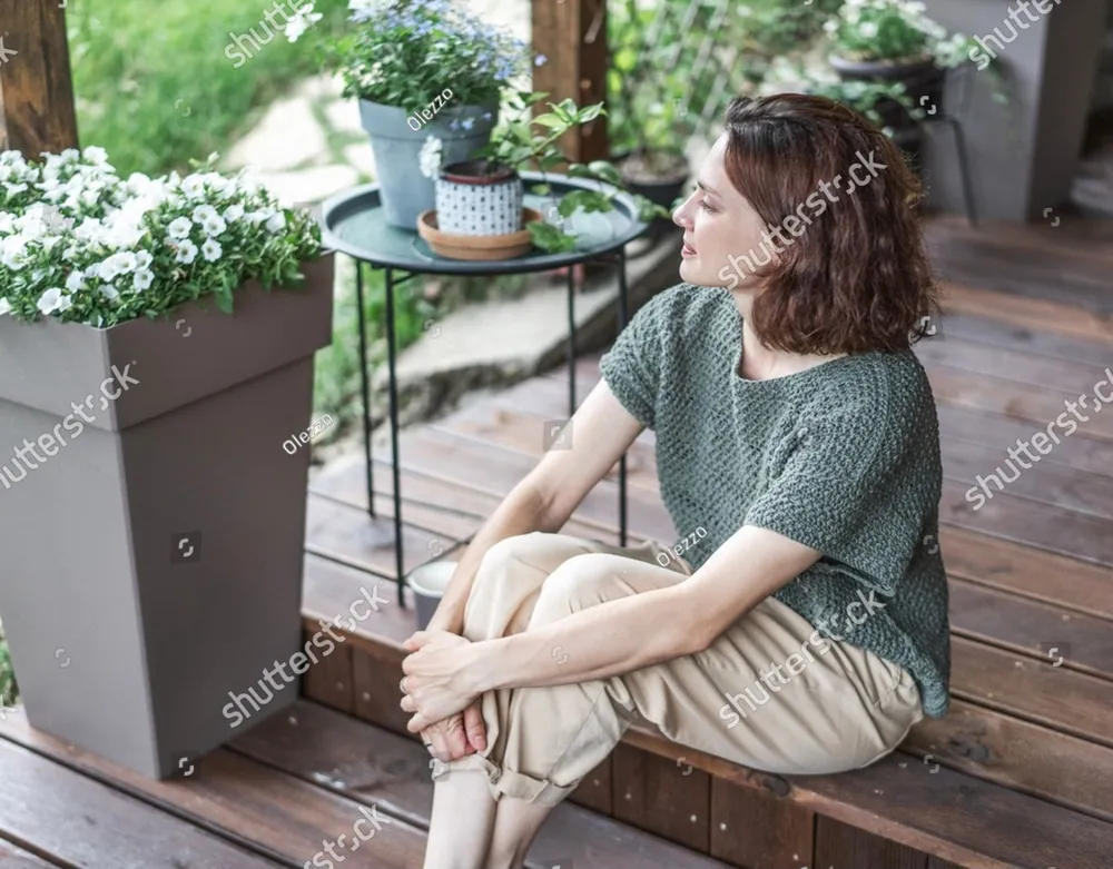 A woman sitting on a porch step with her eyes closed among potted flowers