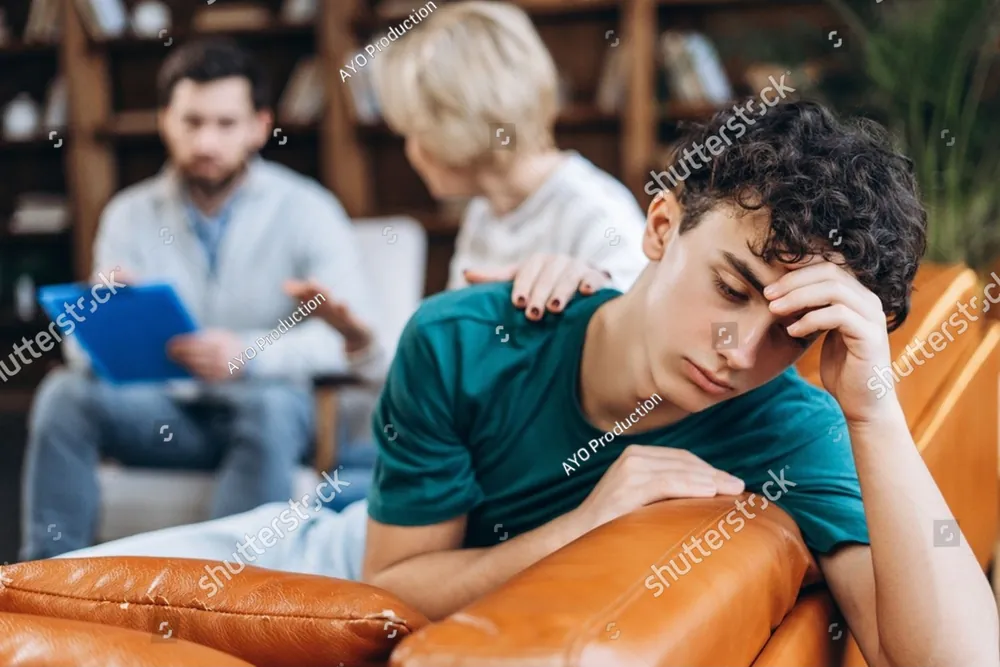 A teenage boy resting his head in his hand on a sofa, his parents behind him
